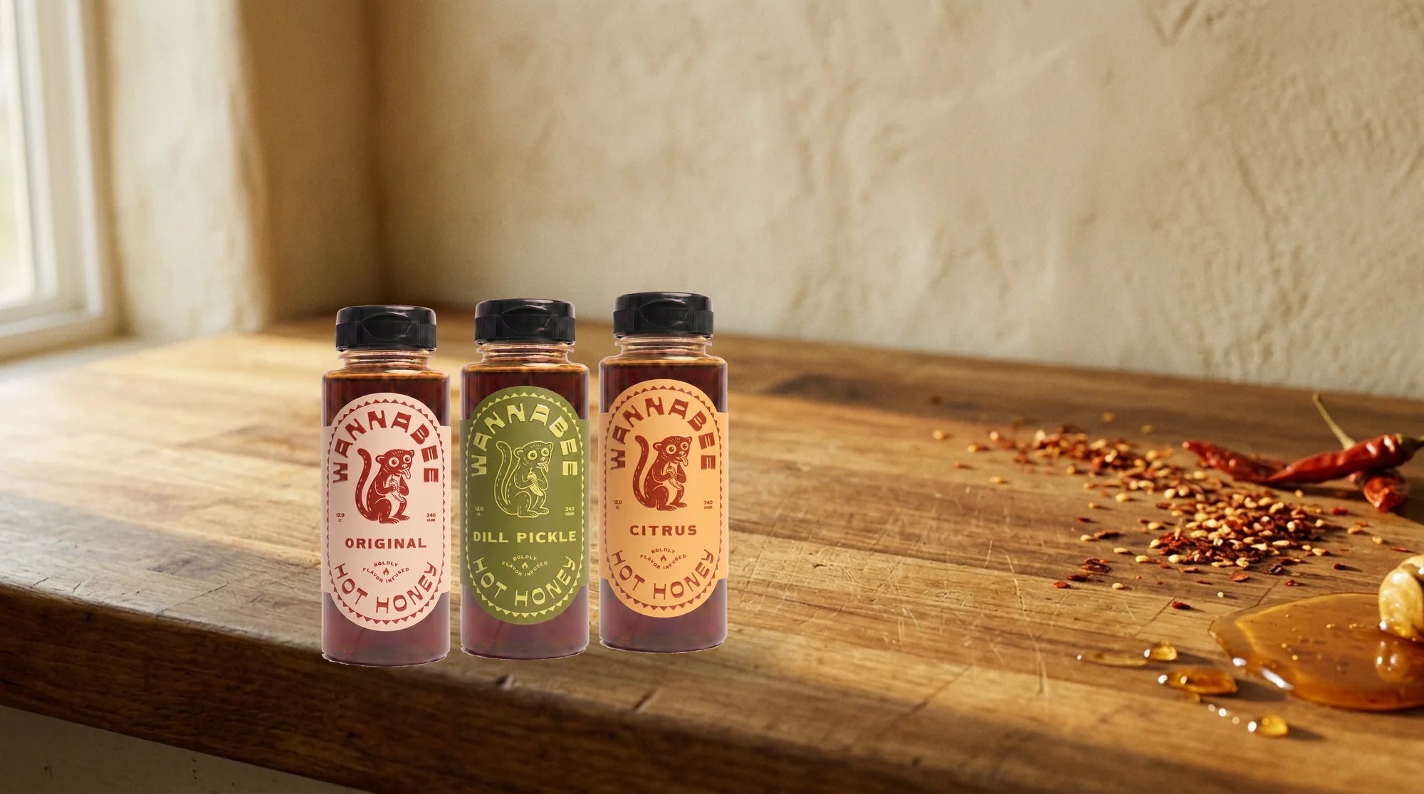 Wide view of three Wannabee hot honey bottles with chili flakes and honey on the counter.