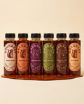 All six Wannabee hot honey bottles standing in a row on a warm cream background — Original, Buffalo, Barbecue, Dill Pickle, Everything Bagel and Citrus — with a pool of amber honey spreading at their base