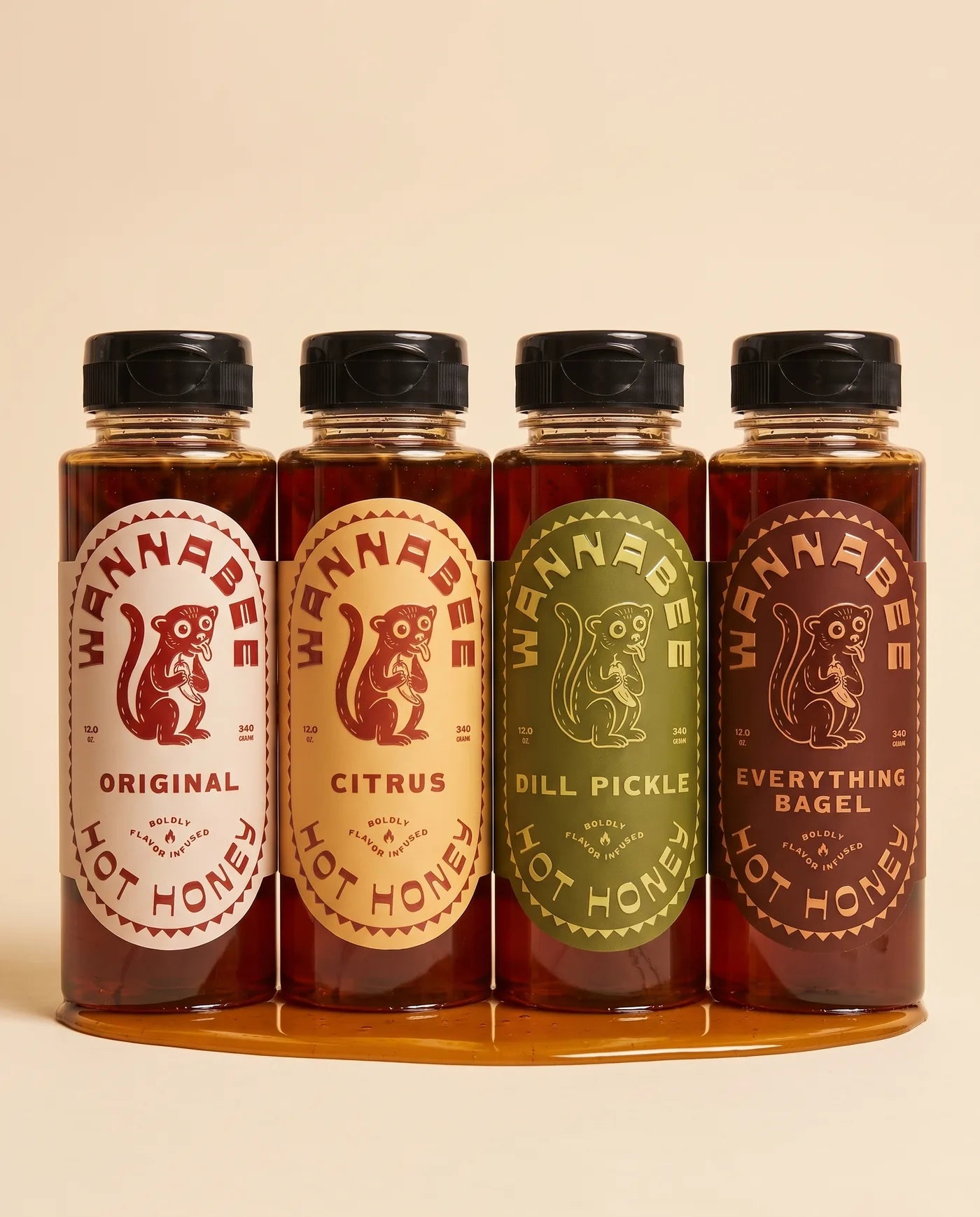 Four Wannabee hot honey bottles standing in a row on a warm cream background — Original, Citrus, Dill Pickle and Everything Bagel — with a pool of amber honey spreading at their base